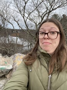 Photo of Amber-Kavka-Warren in a poofy green coat in front of some rocks, barren trees, and water.